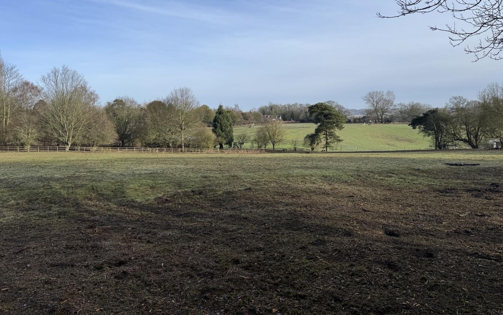 Bearsted__Caring_Lane - SJD Projects Open countryside scene: green pastures with a line of trees and a wooden fence, under a clear blue sky; foreground is muddy and bare.