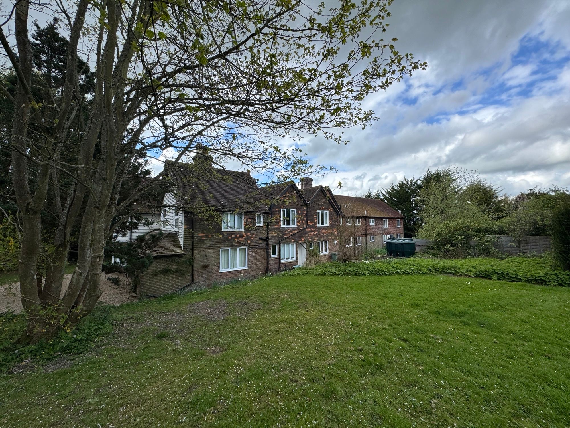 Goudhurst__Old_Poplars - SJD Projects Brick apartment buildings viewed from a grassy yard, with a large tree in the foreground and a cloudy sky above.